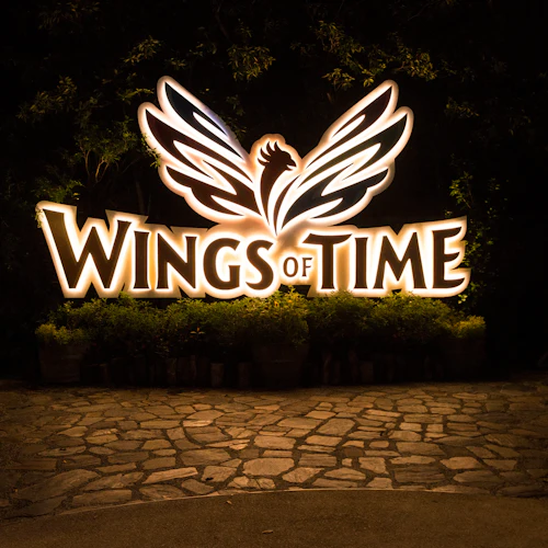 Beleuchtetes "Wings of Time"-Schild mit geflügeltem Logo vor einem dunklen, baumbestandenen Hintergrund mit steingepflastertem Boden.