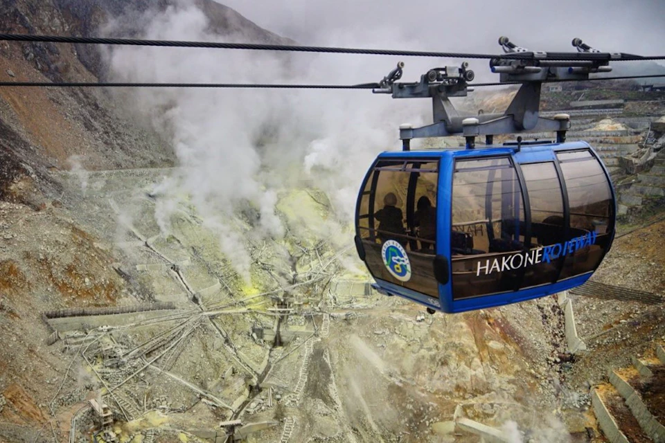 A blue cable car labeled 'Hakone' travels past a steaming volcanic landscape with rugged terrain and visible piping.