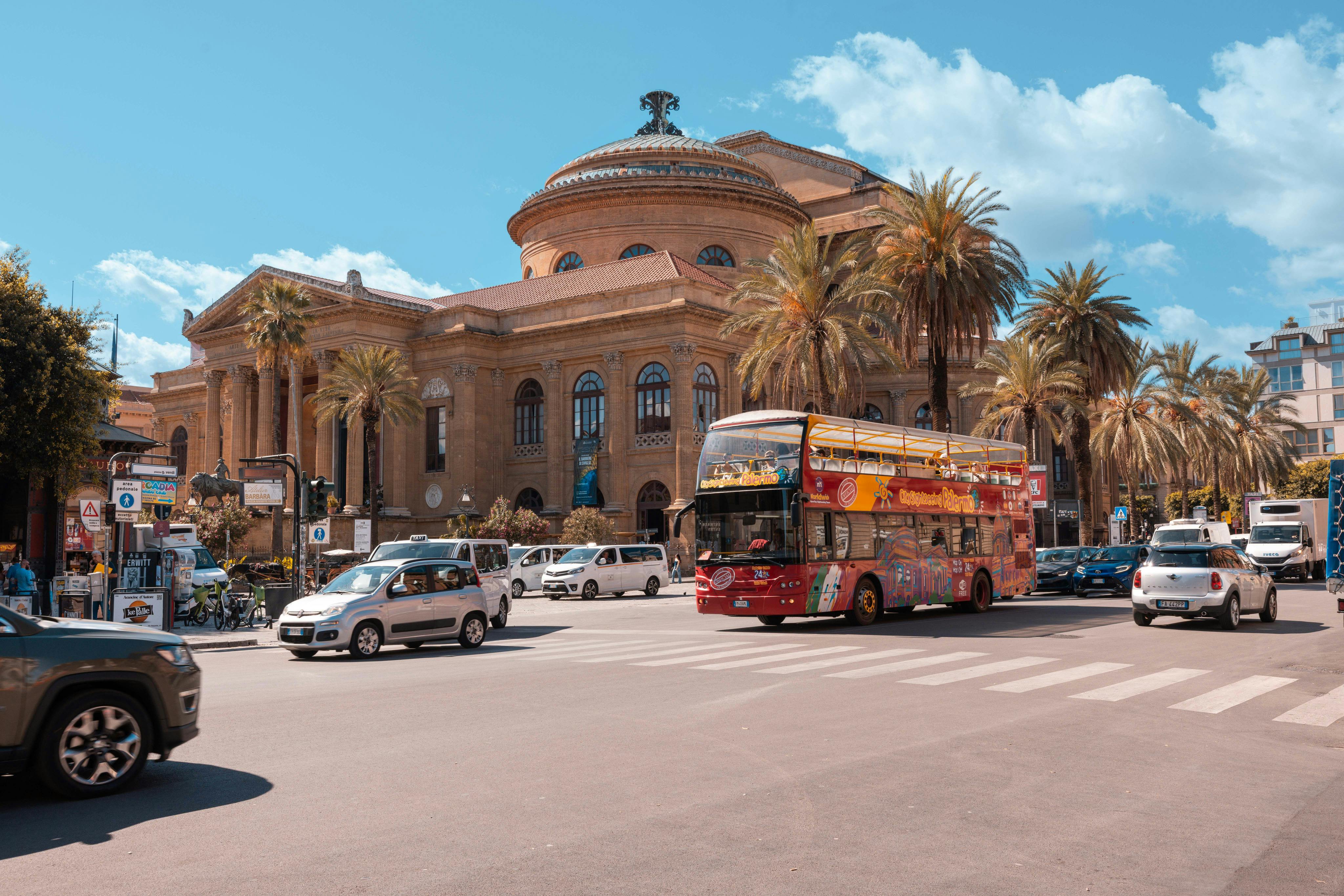 Un autobus turistico rosso a due piani e delle auto davanti a un edificio storico con cupola e palme sotto un cielo azzurro.