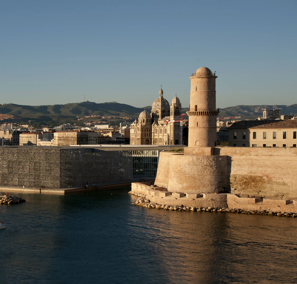 A historic lighthouse and fort by the waterfront with a modern building and mountains in the background under a clear sky.