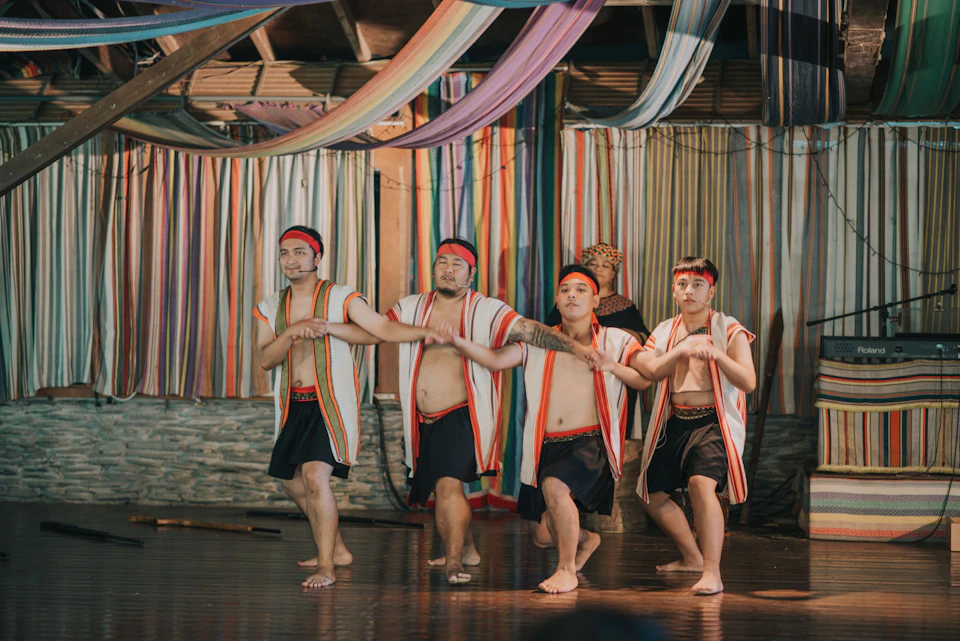 Four men dressed in traditional attire perform a dance on a stage with colorful striped fabric decorations.
