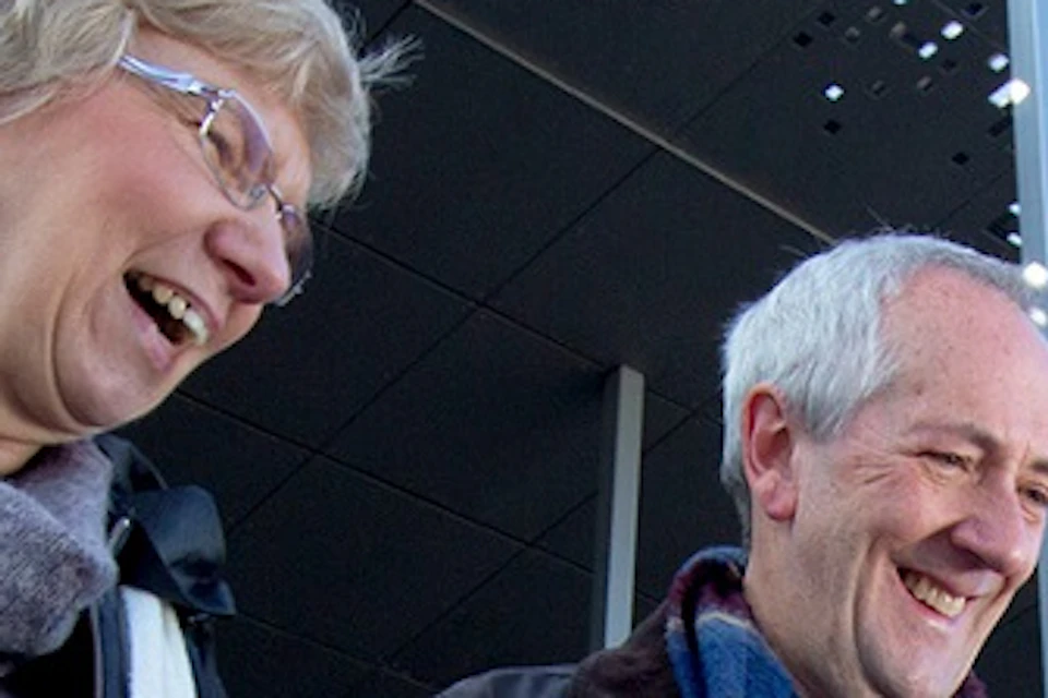 Two elderly people smiling and looking at an outdoor informational display. Modern building with large pillars in the background.