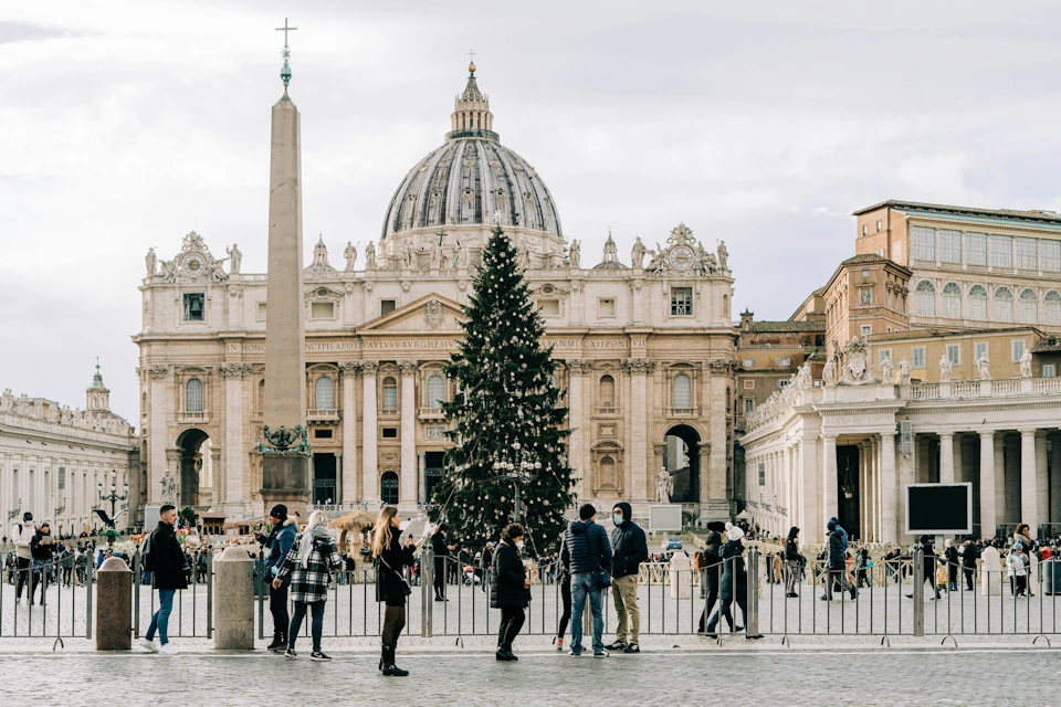 People in winter clothing stand near a decorated Christmas tree and an obelisk in a large square with an ornate building in the background.