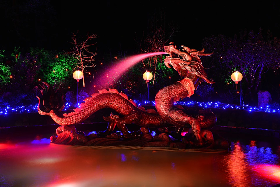 Illuminated dragon statue spouting water at night, surrounded by colorful lights and lanterns against a dark background.