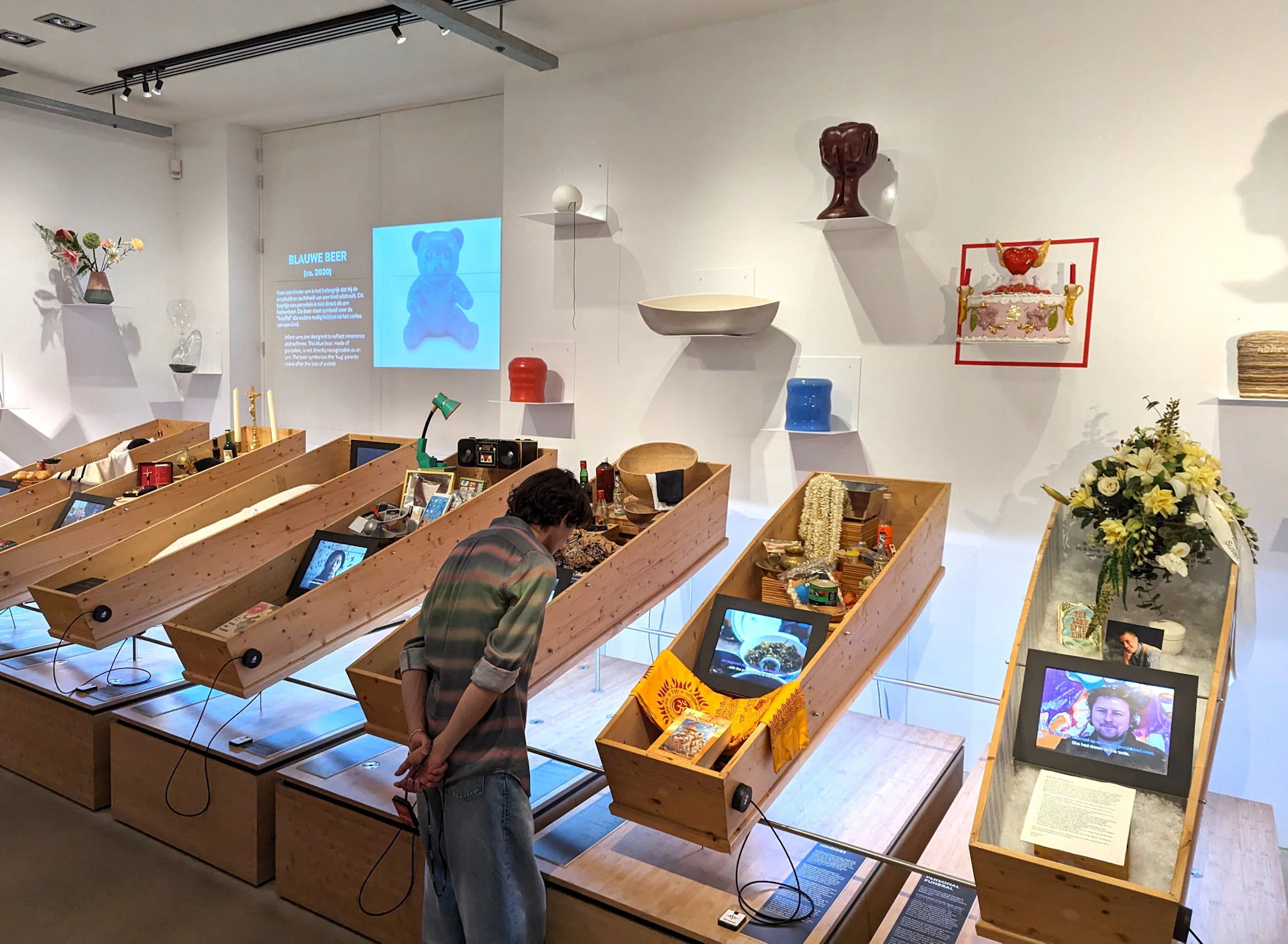 An art exhibition display with wooden angled shelves holding various objects and screens. A person is looking at one of the displays.