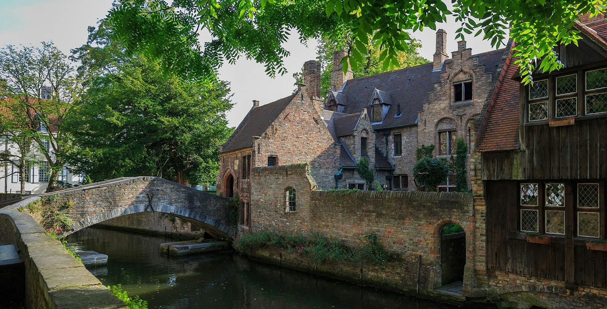 Un bâtiment en briques de style médiéval avec des fenêtres cintrées et un pont en pierre au-dessus d'un canal, entouré d'arbres verts et de feuillage.