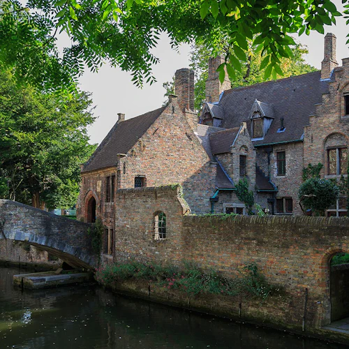 En tegelbyggnad i medeltida stil med välvda fönster och en stenbro över en kanal, omgiven av gröna träd och lövverk.