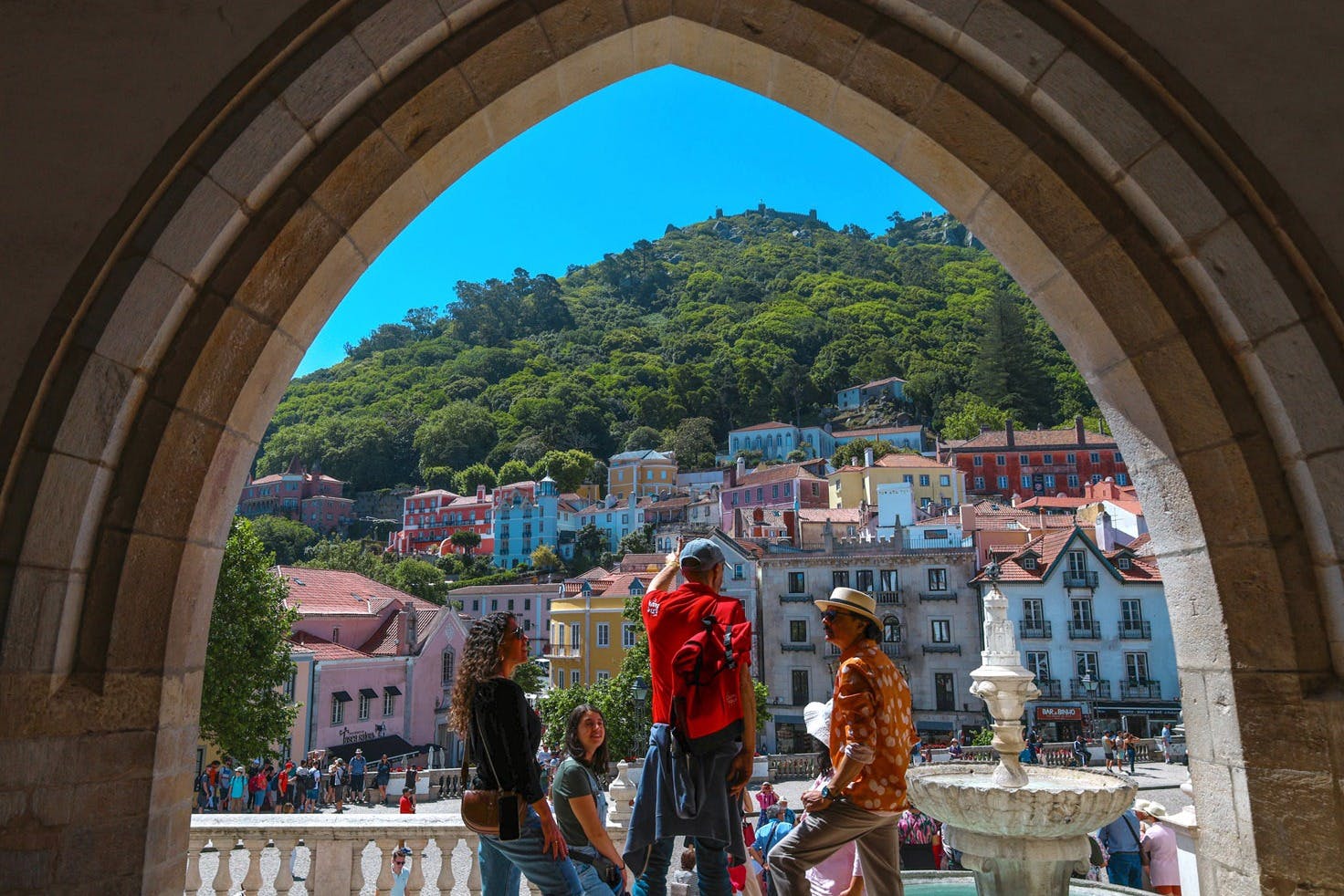 A group of people stands under an archway, facing a hillside with colorful buildings and lush greenery in the background.