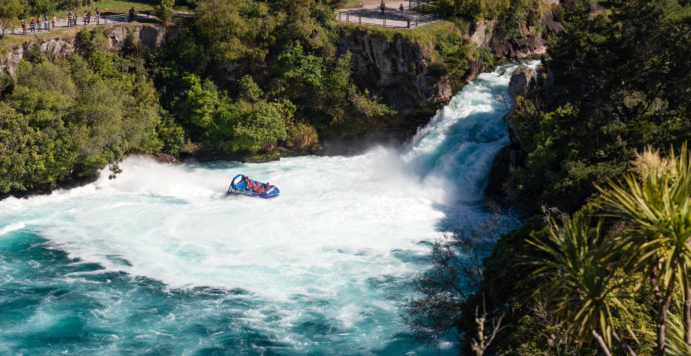 A jet boat navigates white-water rapids in a rocky, forested river gorge under a clear sky.