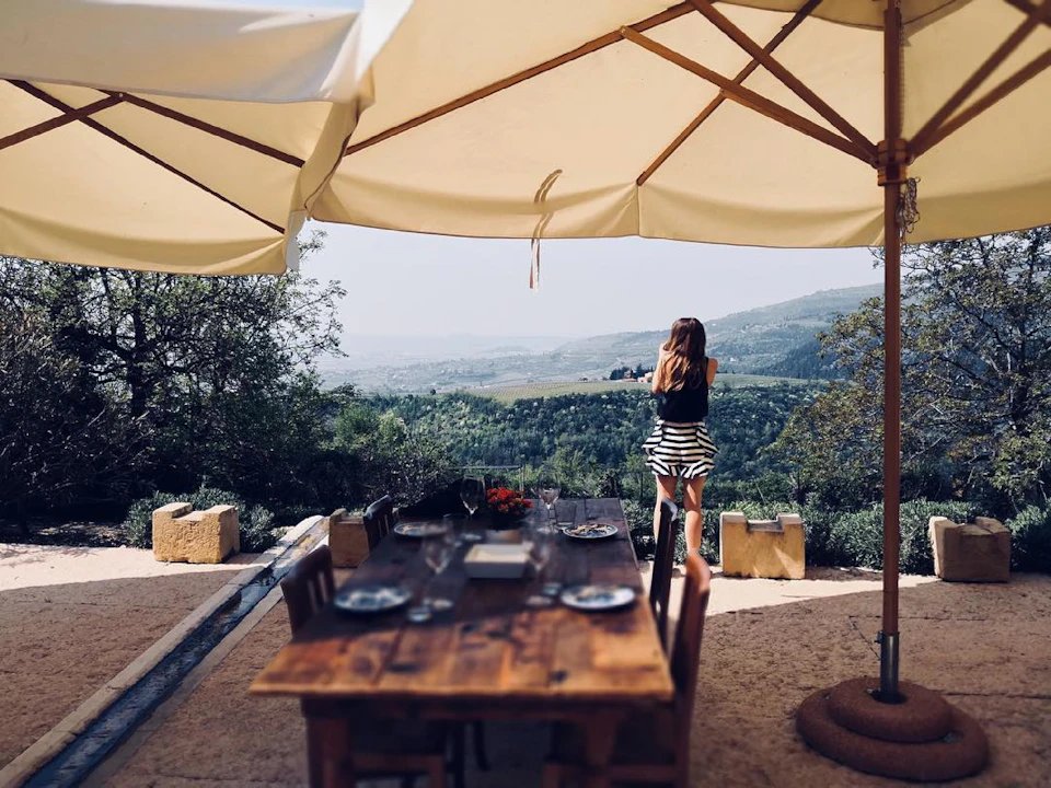 A woman stands near an outdoor dining table under large umbrellas, overlooking a scenic valley and distant hills.