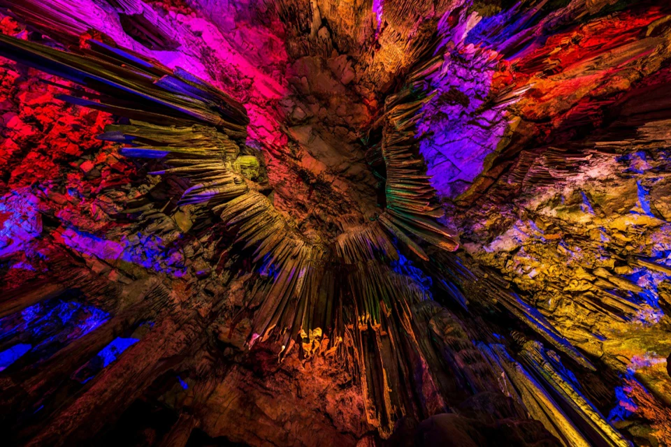 A cave ceiling with colourful lighting highlighting stalactites and rugged rock formations.