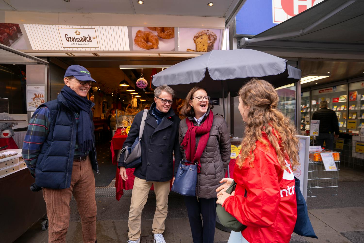 Um grupo de quatro adultos a interagir e a sorrir no exterior de uma padaria, com um guarda-chuva e um letreiro visíveis em segundo plano.
