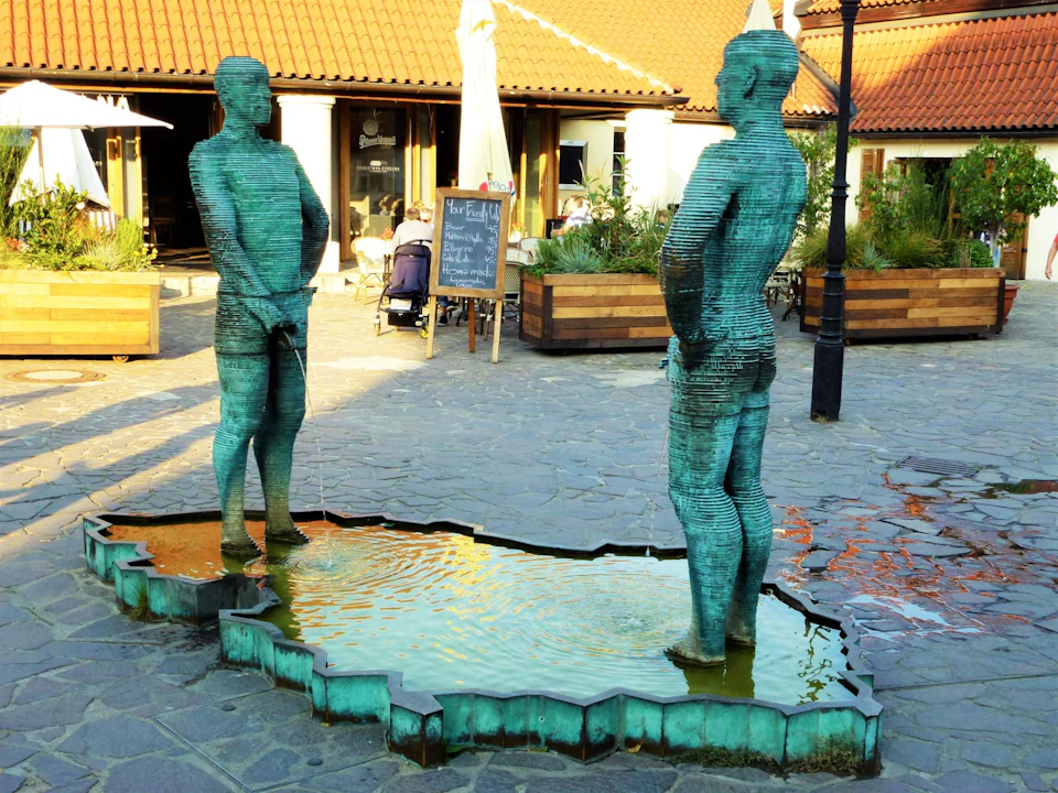 Two metal statues of men stand in shallow water in a courtyard near a building with a tiled roof.