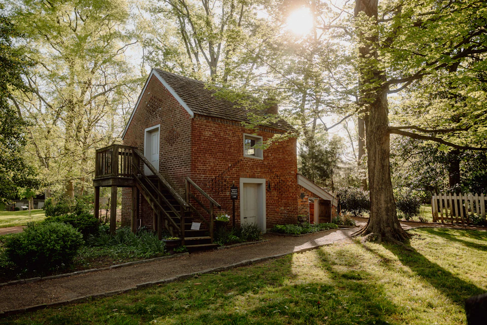 A small brick house with an external staircase, surrounded by trees and greenery, bathed in sunlight.