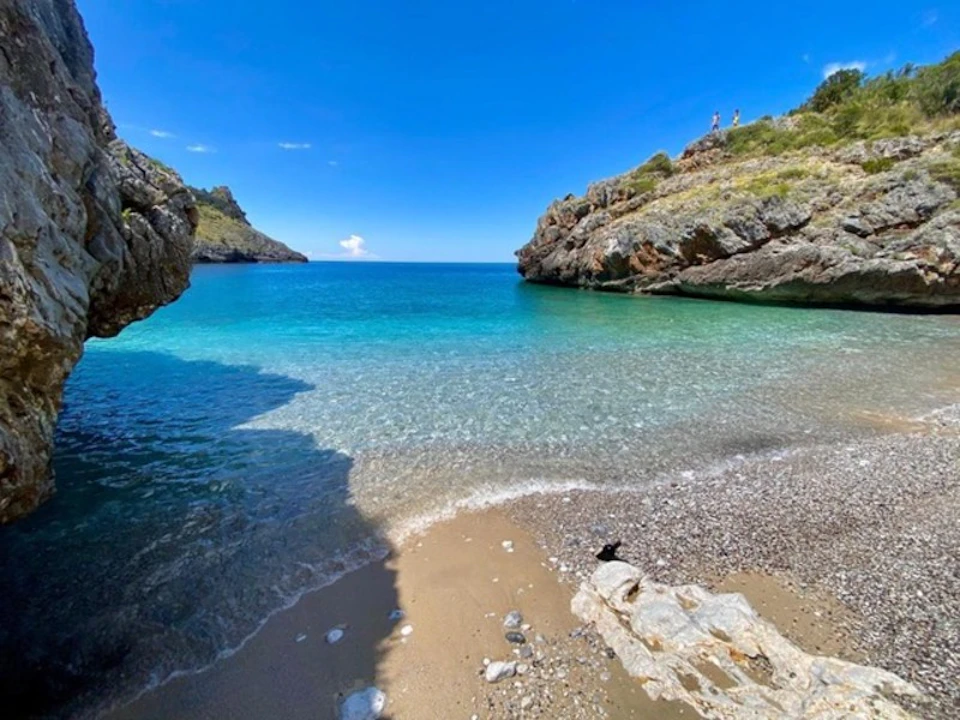 Secluded beach cove with clear blue water, surrounded by rocky cliffs. Small pebbles and sand on the shore under a bright blue sky.