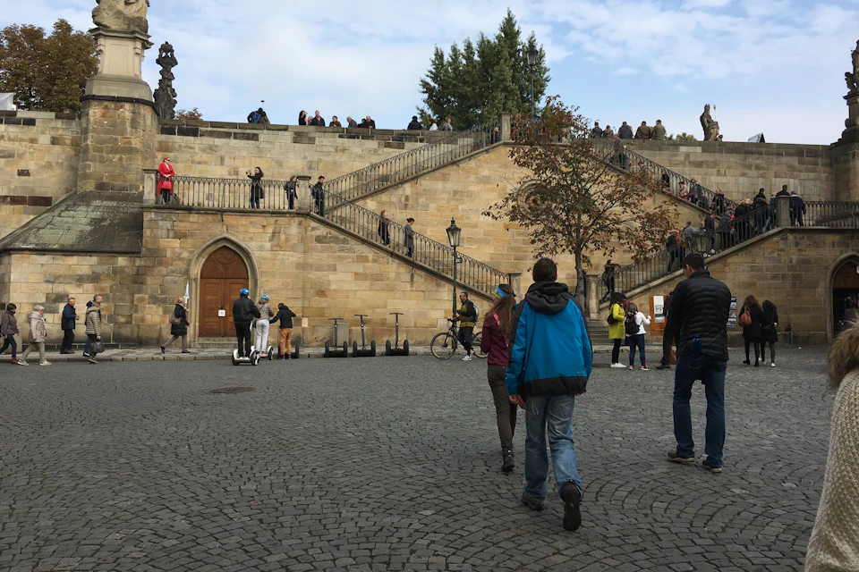 People walking in a cobblestone plaza with a staircase and stone walls in the background, under a partly cloudy sky.