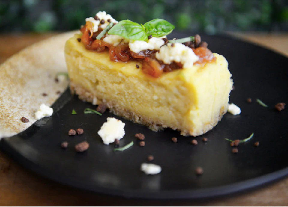 A slice of creamy cheesecake topped with basil, caramelized fruit, and crumbles, served on a black plate.