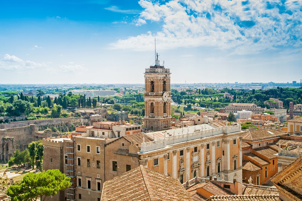 Historic clock tower amid rooftops and green landscape under a partly cloudy sky, with city buildings and greenery in the background.