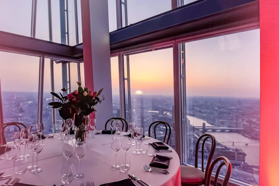 Elegant dining setup near large windows with sunset cityscape view. Table with floral centerpiece, wine glasses, and cutlery.