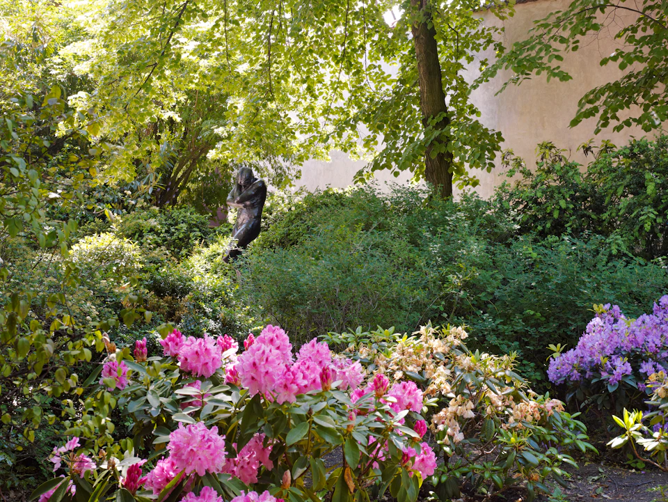 Lush garden with vibrant pink and yellow flowers, dense green foliage, and a dark statue partially hidden among the trees.