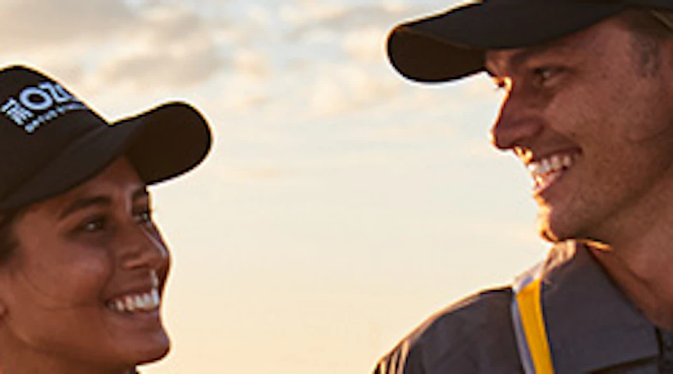 Two people in safety gear and helmets, holding hands, smiling at each other on an elevated platform at sunset.