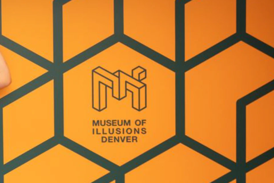 Two people posing in front of an orange wall with a geometric pattern and the text 'Museum of Illusions Denver.'