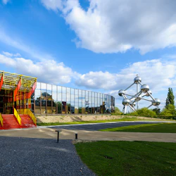 Modern röd-gul struktur bredvid en spegelbyggnad och en metallskulptur, på en grön gräsmatta under en blå himmel med moln.