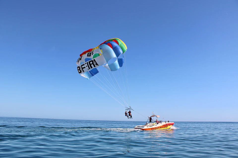 Two people parasailing over a calm ocean, tethered to a colorful parachute attached to a motorboat labeled "Parasailing."