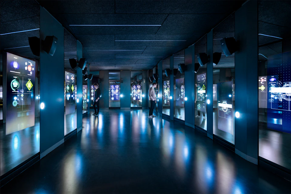 Two people interact with glowing touch screens in a dimly lit, futuristic room with reflective surfaces and ambient blue lighting.