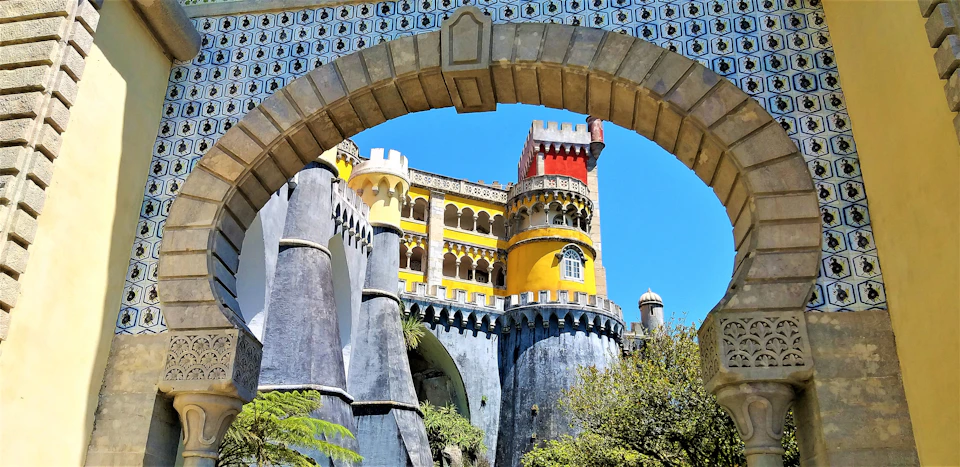 Colorful castle with yellow, red, and gray towers viewed through a decorative stone arch, under a clear blue sky.