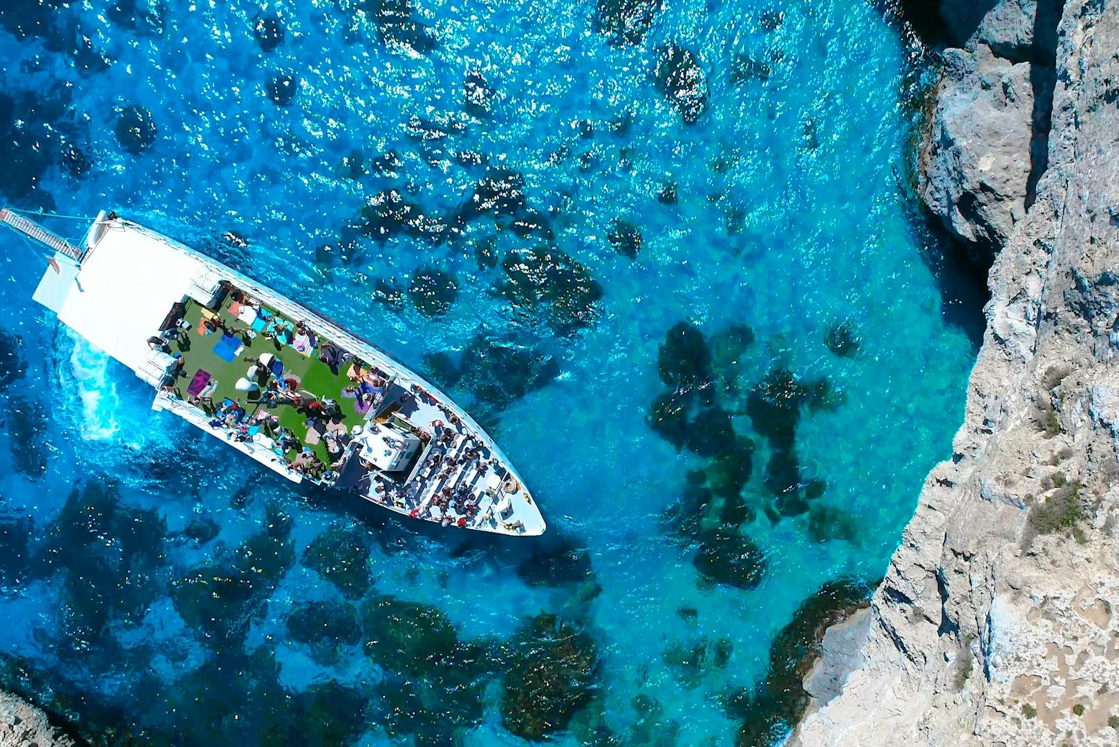 Luftaufnahme eines weißen Bootes mit Menschen an Deck in klarem, blauem Wasser nahe der felsigen Küste.