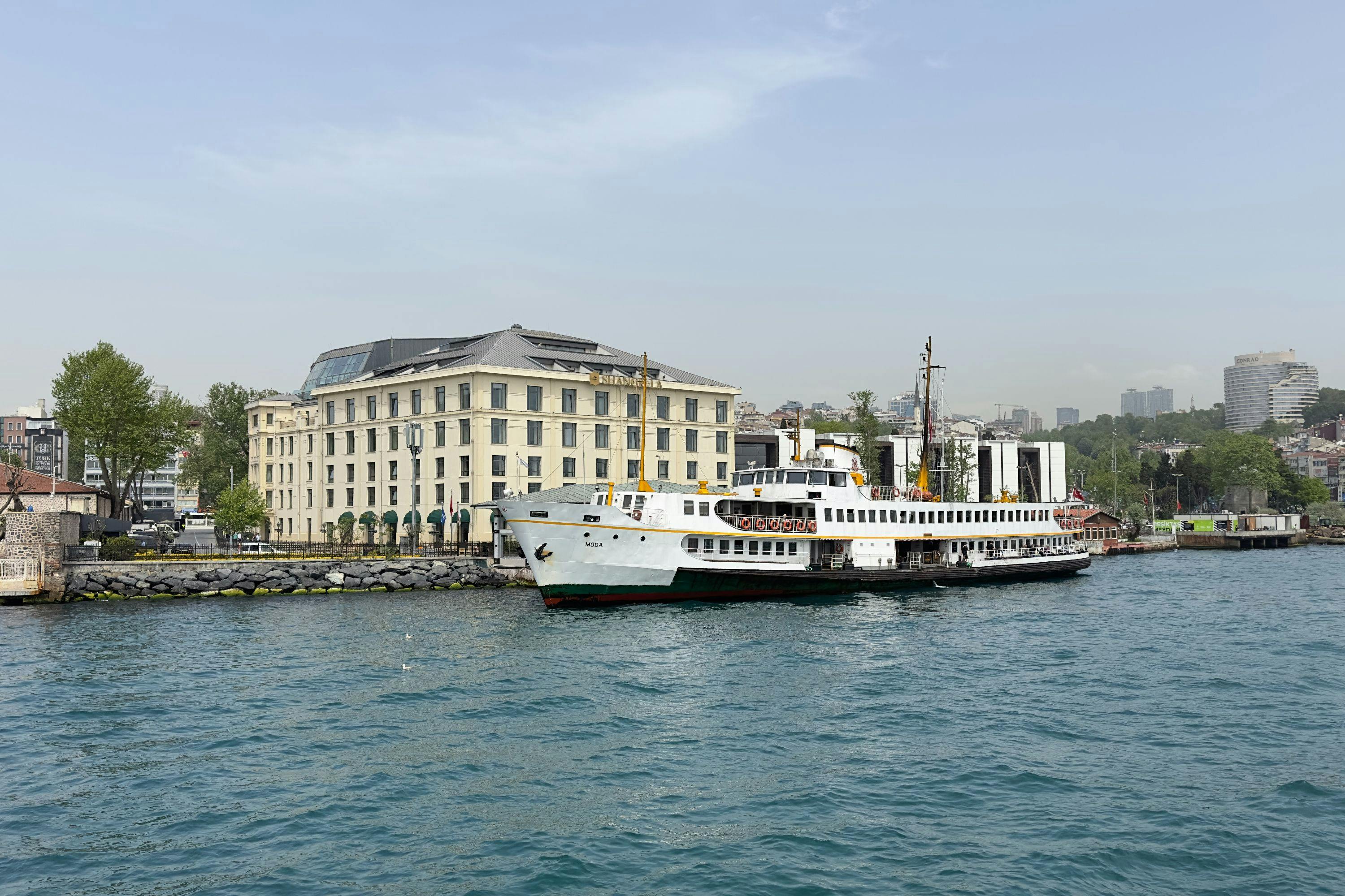 A ferry docked near a waterfront building on a clear day, with calm water and city structures in the background.