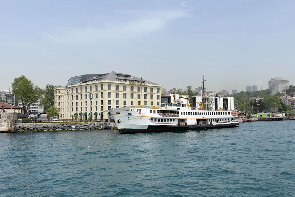 A ferry docked near a waterfront building on a clear day, with calm water and city structures in the background.
