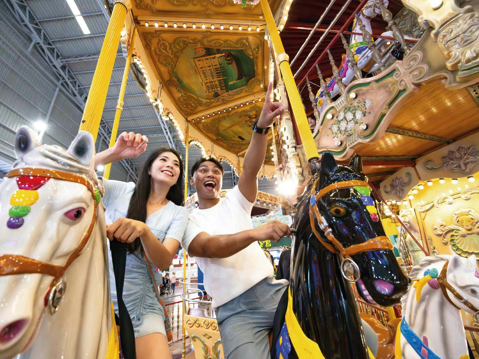 Two people smiling and enjoying a carousel ride with decorative horses in an indoor amusement park.
