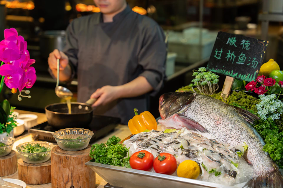 A chef cooking near a display of fresh fish, vegetables, and herbs in a kitchen setting.