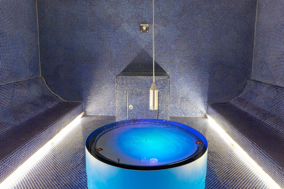 A modern steam room with blue mosaic tiles, a circular illuminated water feature, and minimalist lighting.