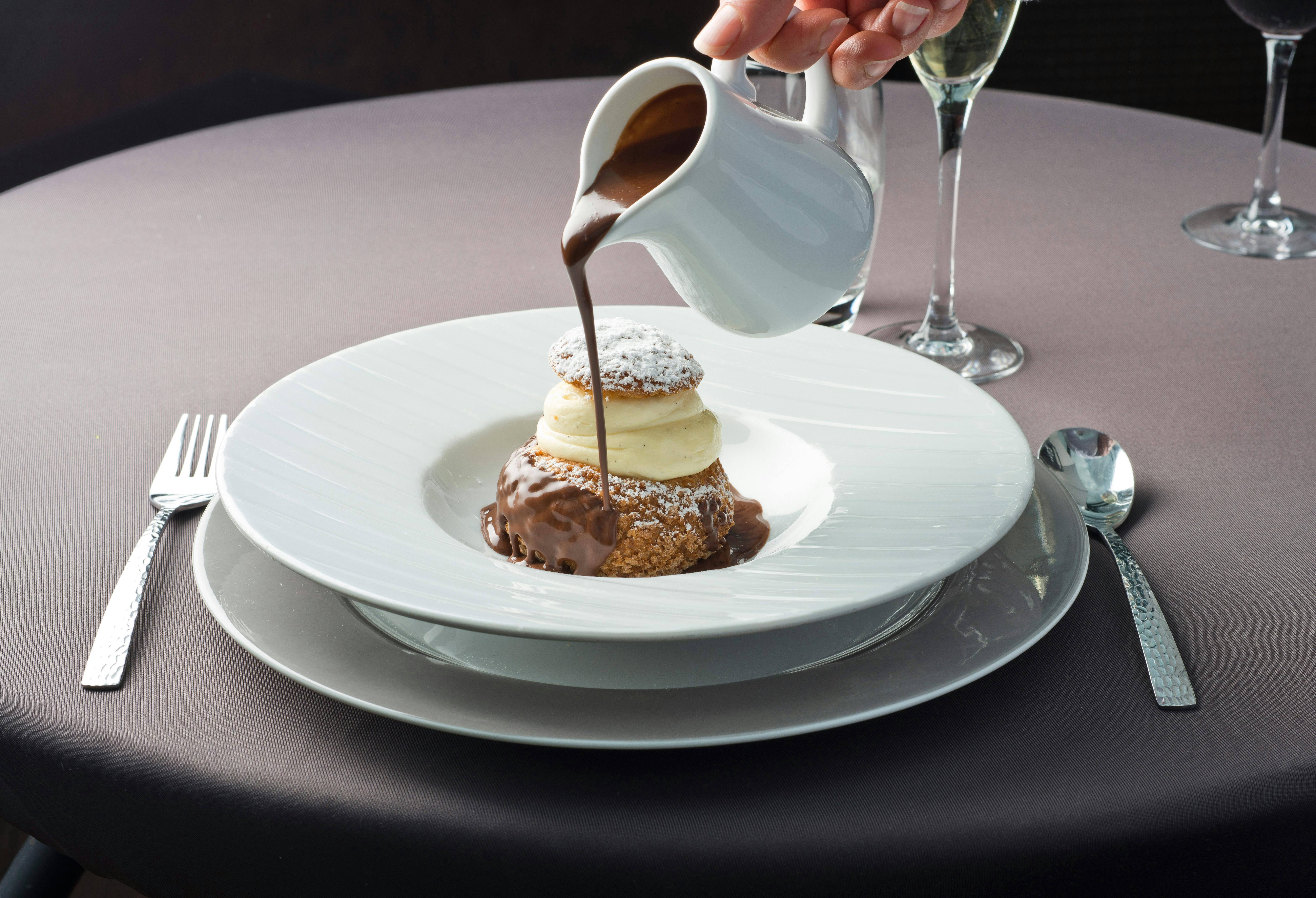 一只手把巧克力酱倒在糕点上，白色盘子上有奶油；附近可以看到一杯葡萄酒和餐具。