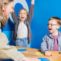 Tre personer spelar ett färgglatt bordsspel, ett barn hejar med armarna lyfta, ett annat barn och en vuxen som ler.