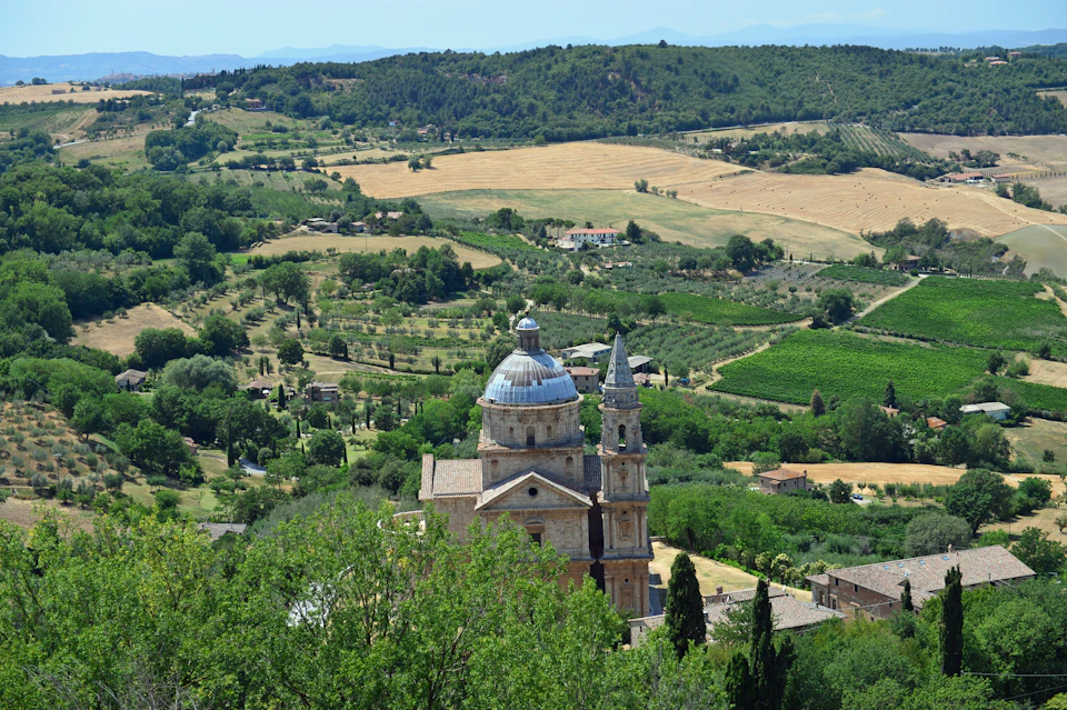 A historic church with a dome and tower is surrounded by lush hills, fields, and scattered buildings under a clear blue sky.