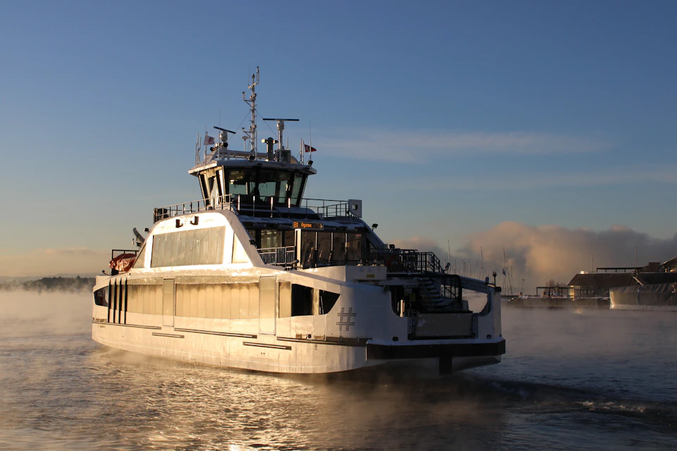 A ferry boat on calm water under a clear sky, with visible steam rising around it, against a harbor backdrop.