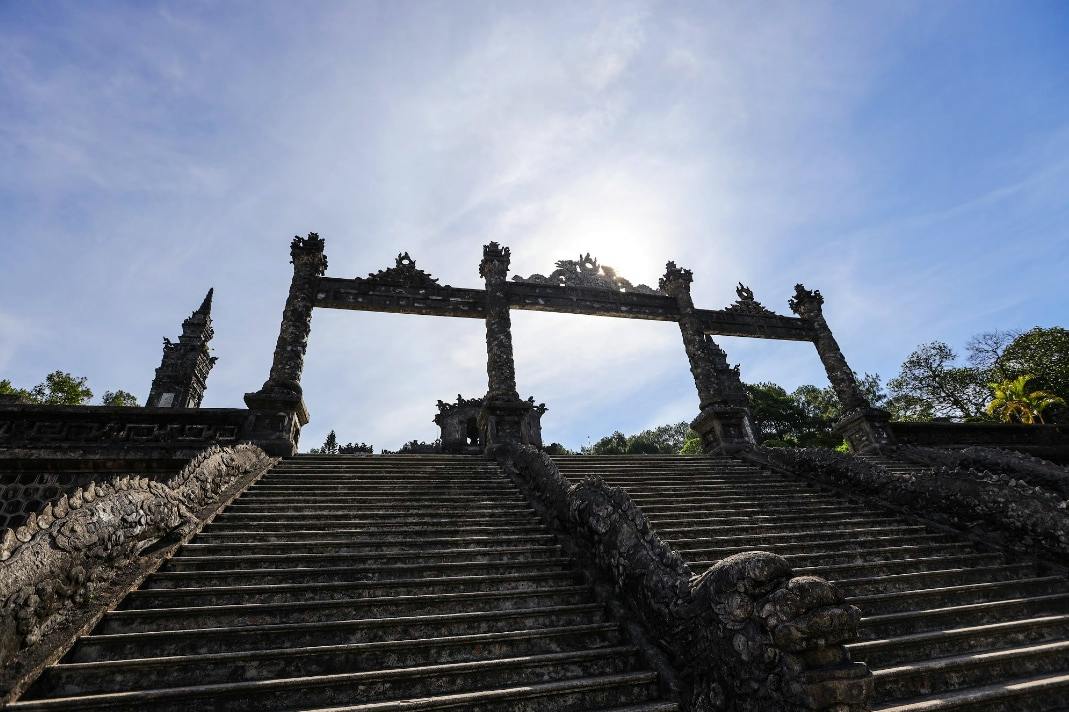 A grand staircase leading to an ornate stone gate under a sunny sky, surrounded by greenery.