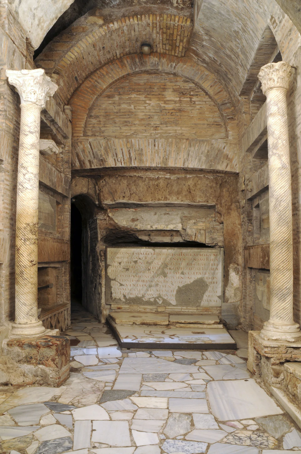 Ancient brick chamber with arched ceiling, two marble columns, niches on side walls, and a large inscribed stone slab at the rear.