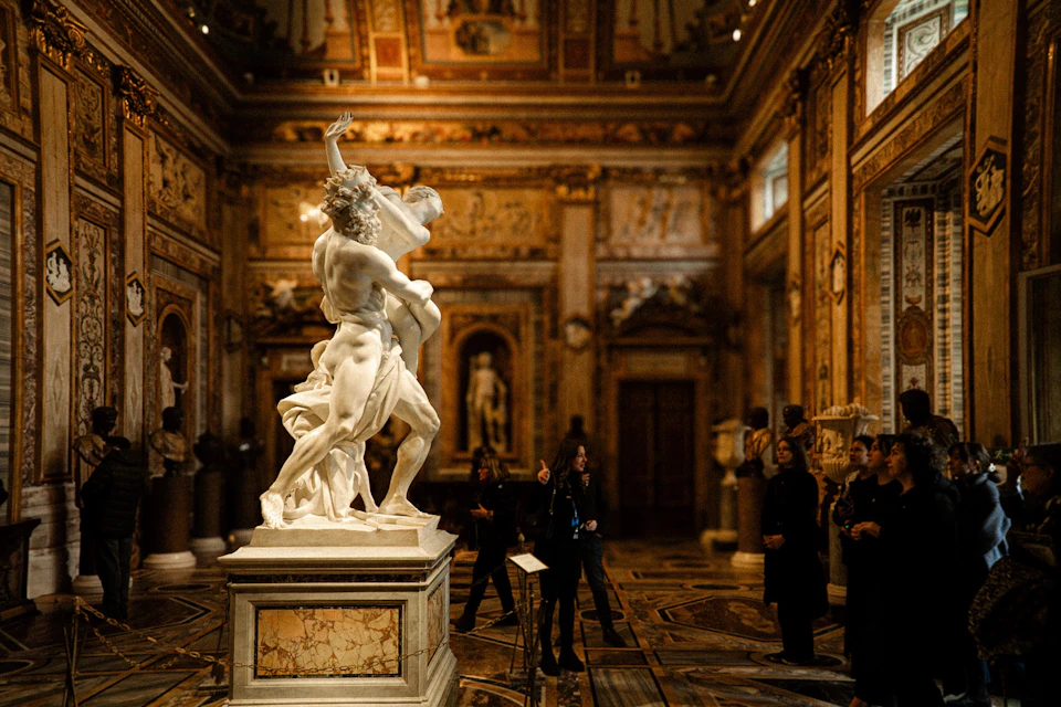 People observing a marble statue of two intertwined figures in an ornate, gold-adorned museum gallery.