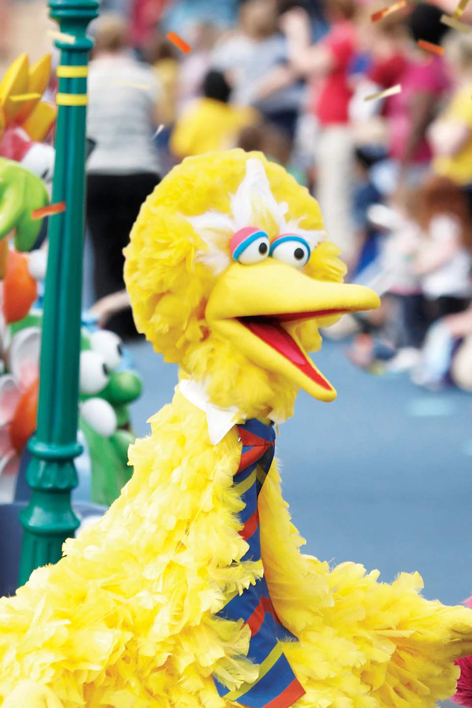 Big Bird, a red Muppet, and two dancers are part of a colorful parade with cheering crowds and festive decorations.