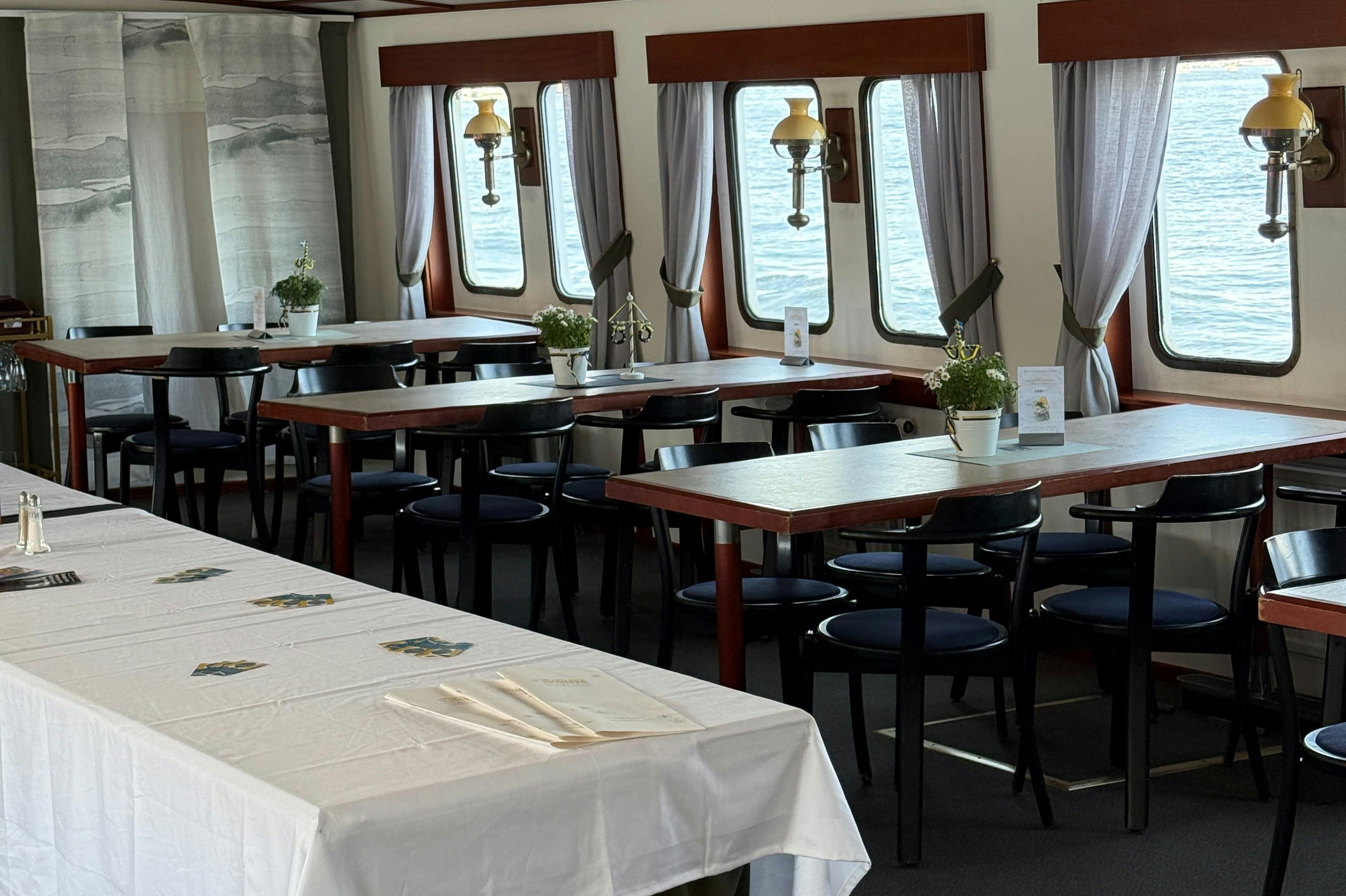 Zona pranzo con più tavoli e sedie su una barca, decorata con piante in vaso, menù e tende alle finestre.