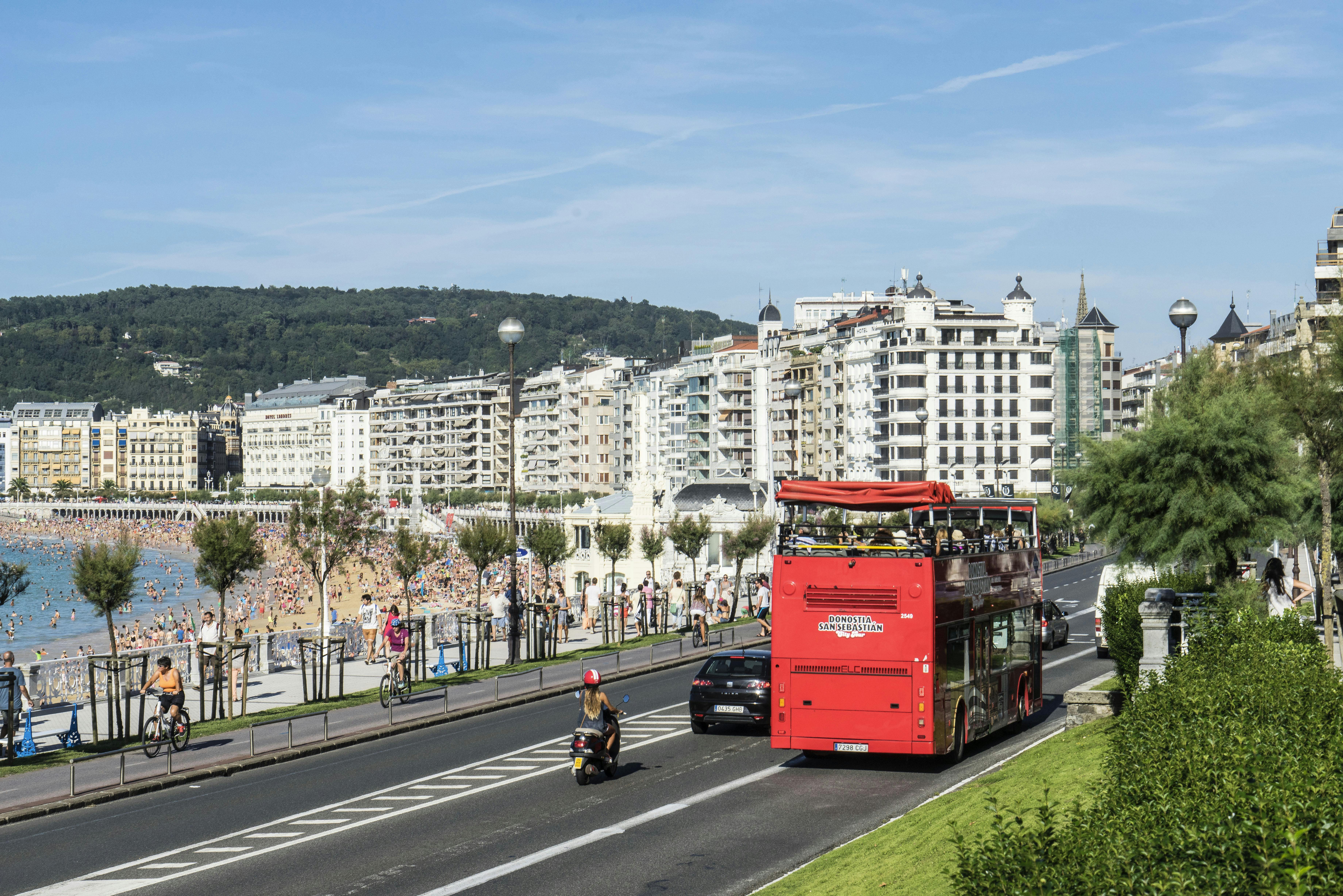 Red double-decker tour bus, motorbike, and car on a road next to a beach with many people and tall buildings in the background.