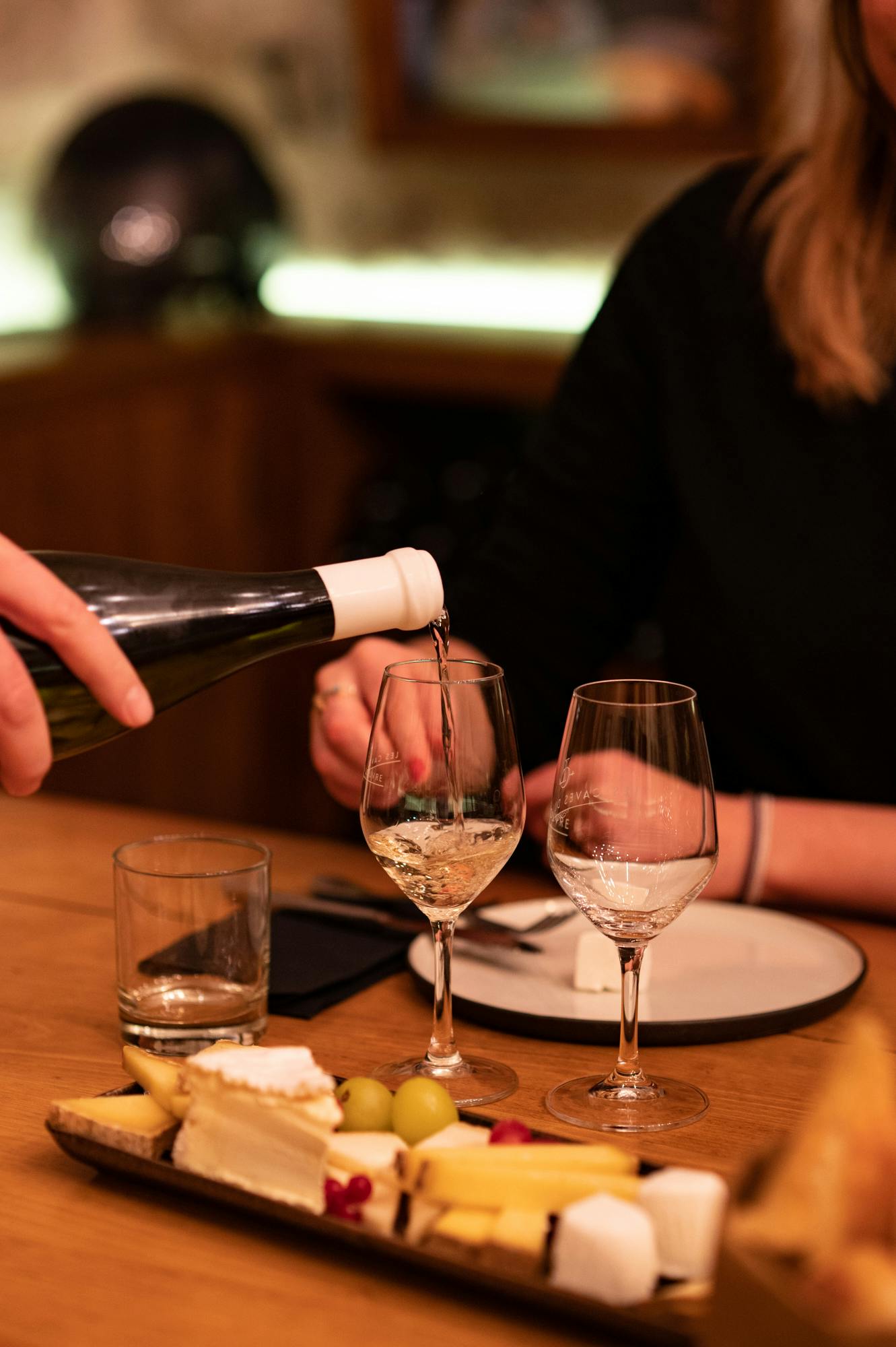 Человек, наливающий белое вино в бокал на обеденном столе с сыром, виноградом и тарелками.