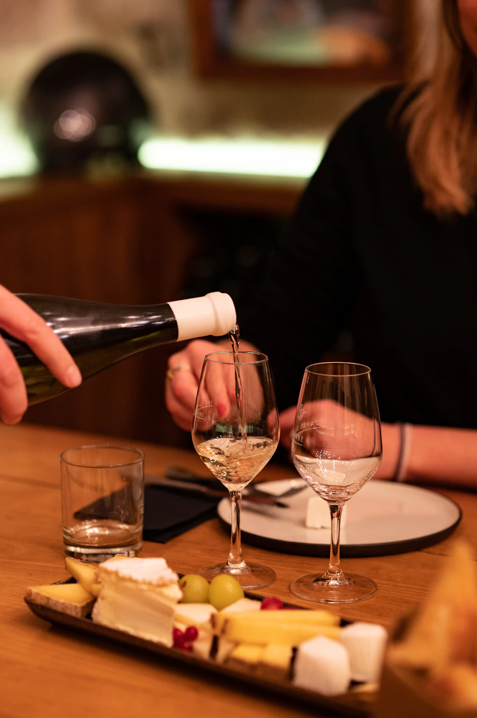 A person pouring white wine into a glass on a dining table with cheese, grapes, and plates.