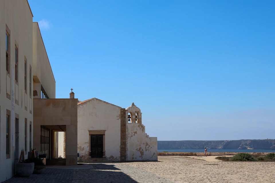 A small white chapel with a bell tower on a bright day, situated near a cobblestone area and overlooking a distant sea.