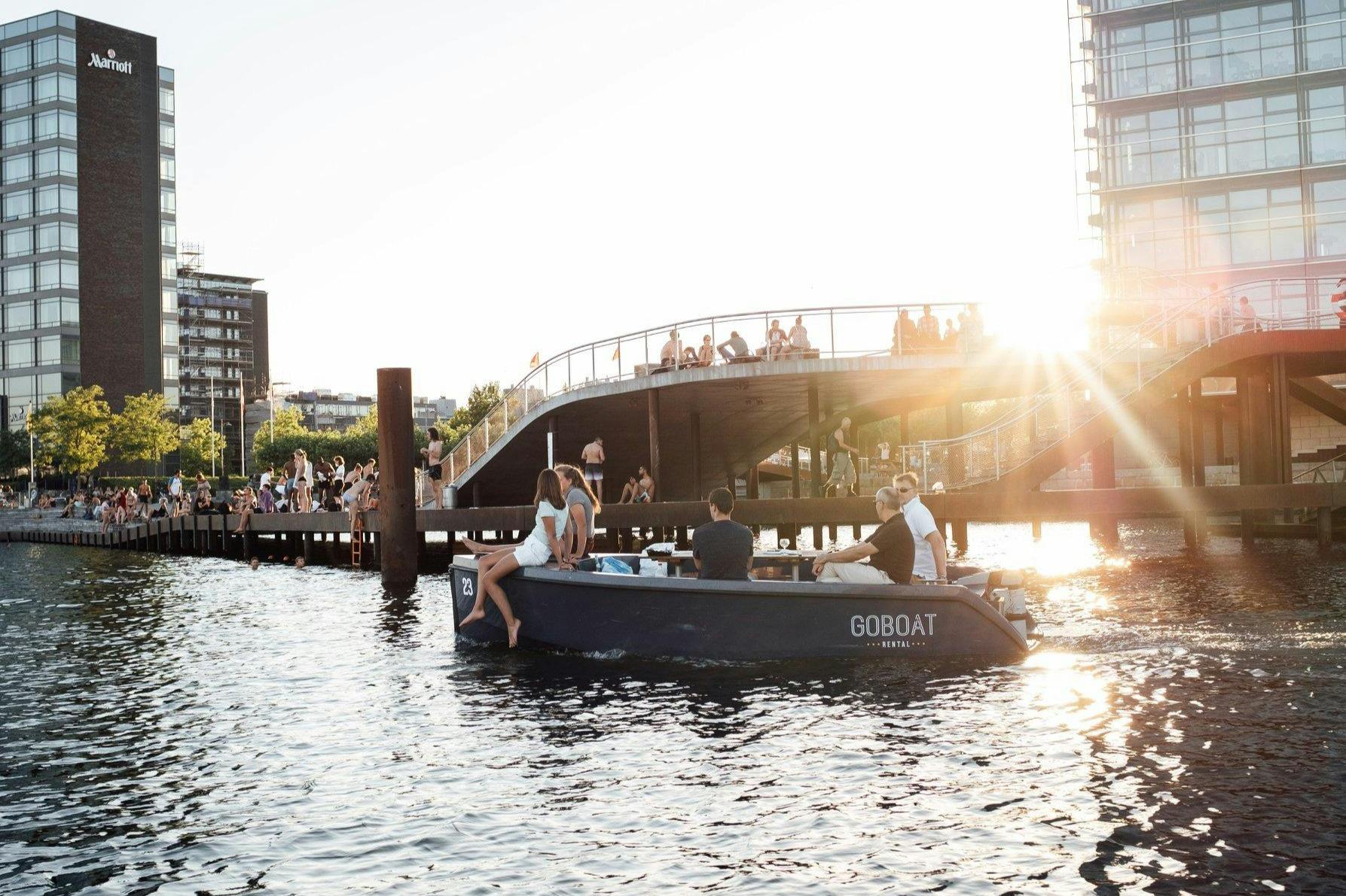 People enjoying a boat ride and others walking on a footbridge over a sunny waterfront with modern buildings in the background.